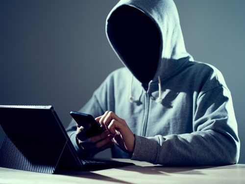 パーカーのフードを被り、顔は影で見えない男がノートパソコンとスマホを捜査している